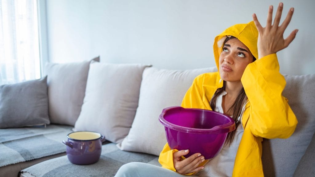 Woman in yellow raincoat holding a bucket to catch a roof leak in her living room.