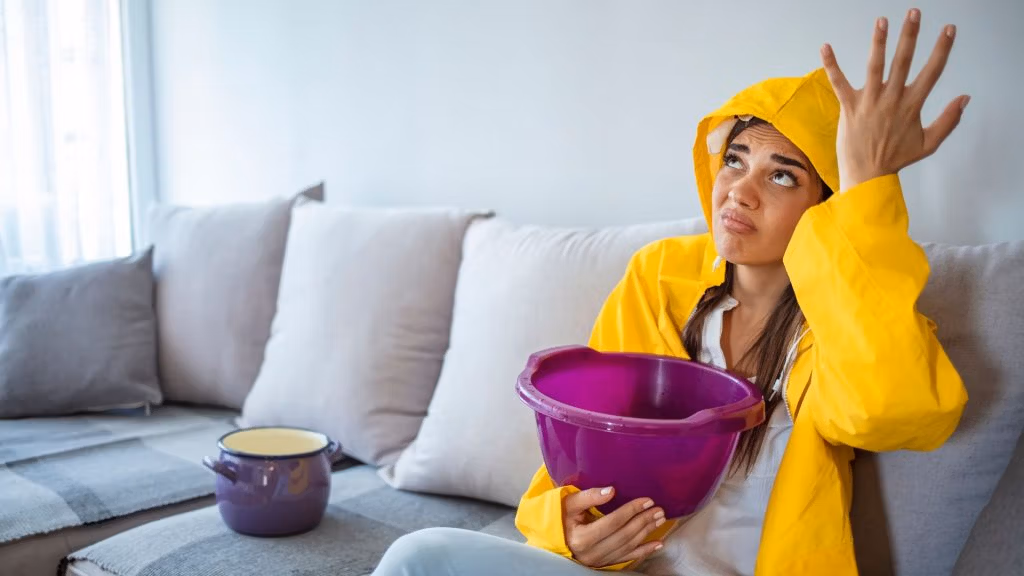 Woman in yellow raincoat holding a bucket to catch a roof leak in her living room.